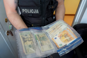 Fotografia kolorowa. Na zdjęciu widoczny policjant w umundurowaniu w kolorze czarnym. Widać sylwetkę policjanta poniżej głowy. W rękach policjant trzyma foliowe worki z banknotami polskich złotych oraz w  obcej walucie.