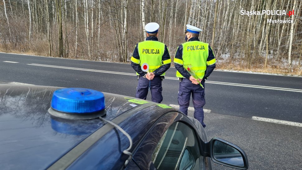 na pierwszym planie nieoznakowany radiowóz z sygnałem świetlnym na dachu, w tle dwaj policjanci, z których jeden trzyma tarcze do zatrzymywania pojazdów a drugi tulipana