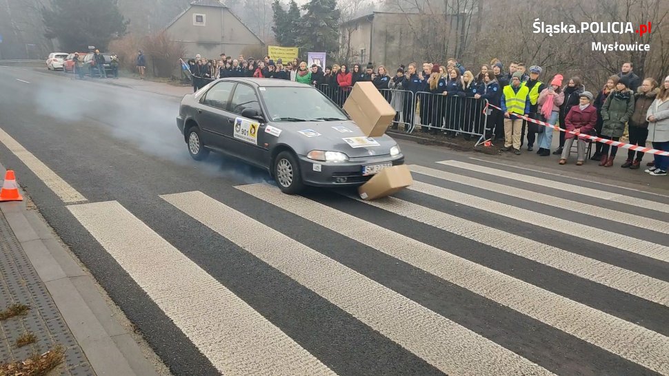 samochód hamujący przed przejściem dla pieszych. Z pod kół widoczny dym. W tle uczniowie przyglądajacy się pokazowi Prędkość i hamowanie