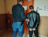 nieumundurowany policjant z zatrzymanym mężczyzną, który ma założone kajdanki na ręce trzymane z tyłu.