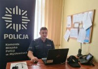 policjant siedzący przy laptopie podczas spotkania w formie online z uczniami. W tle rollup Komendy Miejskiej Policji w Mysłowicach