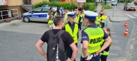 policjanci i dziewczynka rozmawiają z pieszym o zmianach w przepisach. W tle policyjny radiowóz i grupa pozostałych uczniów.