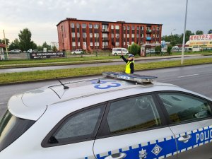 Na pierwszym planie radiowóz, w tle policjantka ruchu drogowego z uniesioną w górę prawą ręką.