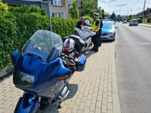 Na pierwszym planie niebieski motocykl. W tle policjanci przeprowadzają badanie stanu trzeźwości kierującego. Za nimi stoi zaparkowany policyjny radiowóz.