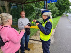 policjantka wręcza elementy odblaskowe kobiecie i mężczyźnie na przystanku autobusowym