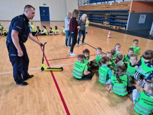 policjant trzyma hulajnogę, przed nim siedzi grupa przedszkolaków w zielonych kamizelkach