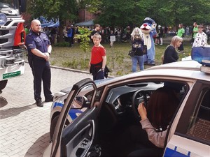 policjant na festynie rodzinnym, w tle widać maskotkę Śląskiej Policji Sznupka. na pierwszym planie dziecko, które siedzi w radiowozie