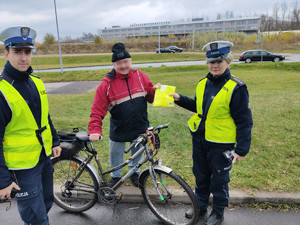 Na zdjęciu znajdują się policjanci wydziału ruchu drogowego, wręczający rowerzyście kamizelkę odblaskową.