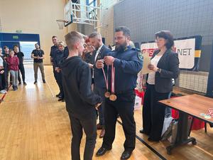 Na zdjęciu znajdują się zwycięscy trzeciego miejsca w turnieju. Komendant Straży Miejskiej wręcza medal uczniowi szkoły podstawowej.