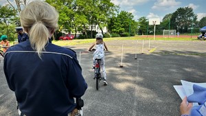 zdjęcie przedstawia dziecko poruszające się na rowerze pokonujące slalom, dziecku przygląda się policjant