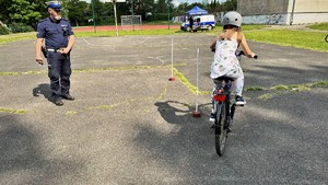 zdjęcie przedstawia dziecko poruszające się na rowerze pokonujące slalom, dziecku przygląda się policjant