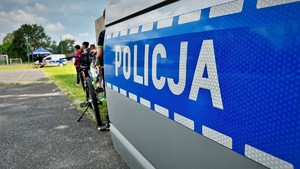 zdjęcie przedstawia zbliżenie na napis POLICJA na radiowozie, w tle widać dziecko trzymające rower