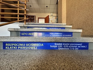 zdjęcie przedstawia naklejkę na schodach z napisem Rozpocznij uciśnięcia klatki piersiowej Środek klatki piersiowej! Tempo 100-120 uciśnięć na minutę! Głębokość 5-6 centymetrów!