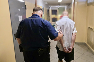 zdjęcie przedstawia zatrzymanego prowadzonego w kajdankach przez policjanta